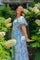 woman wearing a blue and white floral maxi dress, smiling and standing among white flowers in a garden