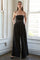 woman wearing a black strapless jumpsuit with a tied waist and wide-leg pants, standing against a bright window