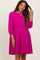 woman wearing a vibrant pink maternity dress with long puff sleeves and a relaxed fit, smiling and touching her hair
