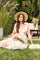 woman sitting on grass wearing a floral maxi dress with pink bow straps and a straw hat, holding a bouquet of flowers