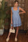 woman wearing a light blue maternity dress with tie shoulder straps and lace trim, holding a beige bag, standing by a plant