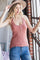 woman wearing a rust-colored ribbed tank top with buttons and blue jeans, standing by a window