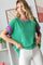 woman wearing a green short sleeve knitted top with purple and pink accents, styled with white jeans