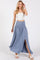 woman wearing a light blue maxi skirt with a front slit and a white cropped top, standing with one hand raised