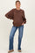 woman wearing a brown oversized maternity sweatshirt and flared jeans, smiling with hands on her hips and a visible baby bump