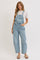 woman wearing light blue denim overalls with a white top, standing with hands in pockets on a neutral background