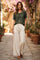 woman wearing a dark green short sleeve top and cream wide-leg trousers, walking down a cobblestone path surrounded by plants