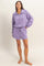 woman wearing a purple zip-up pullover and matching shorts, standing barefoot against a light background