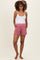 woman wearing a white tank top and pink maternity shorts with pockets, standing barefoot and smiling