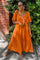 woman wearing an orange tie-front maxi dress with short sleeves and brown flats, standing outdoors with greenery in the background