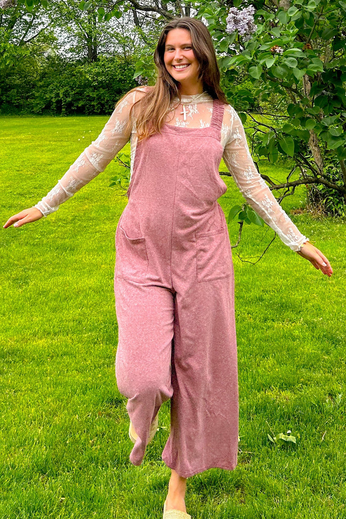 woman wearing a pink linen jumpsuit with wide legs and pockets, paired with a sheer white long sleeve top, smiling outdoors
