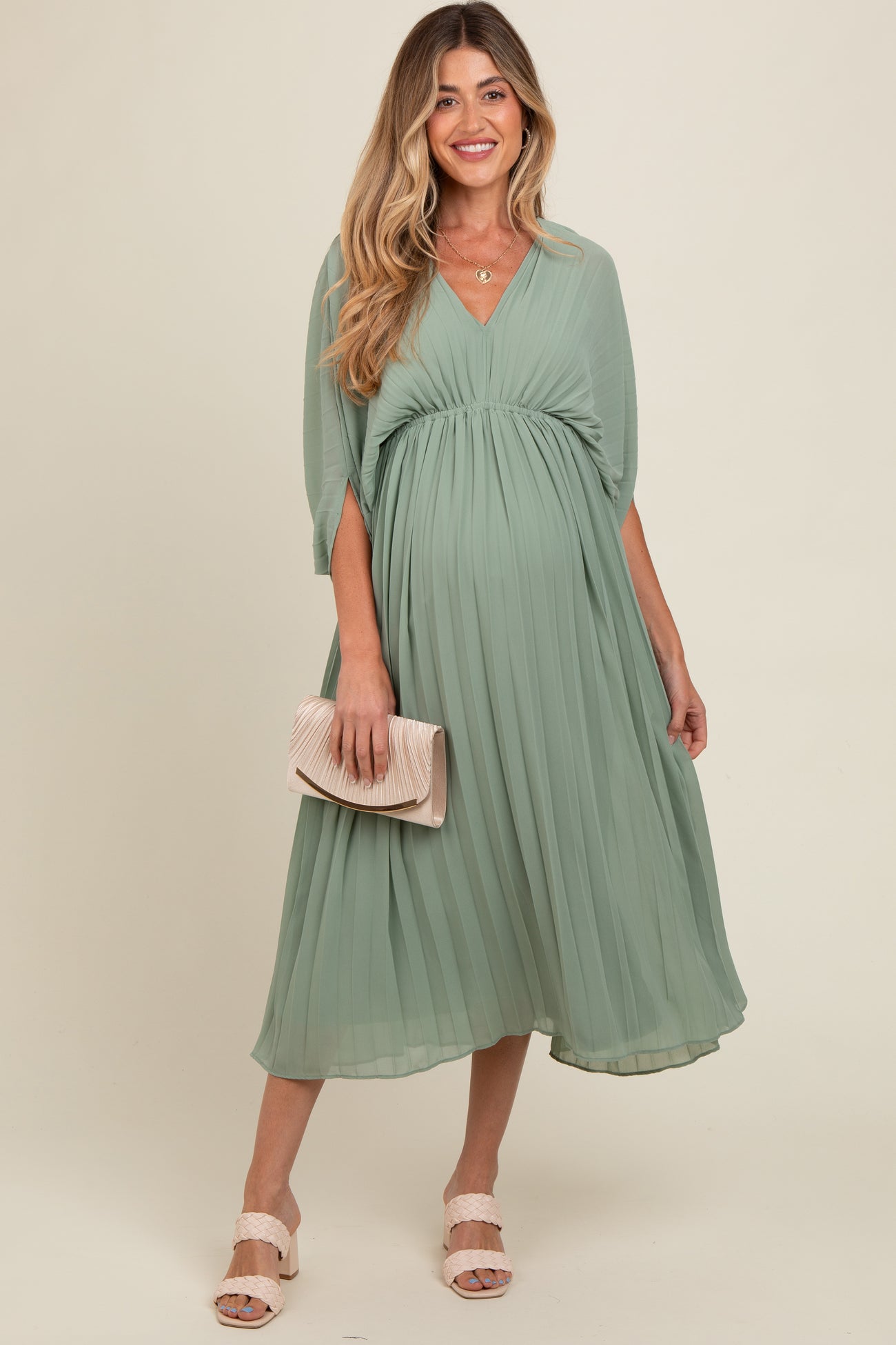 woman wearing a mint green pleated maternity dress with a v-neck and flowing sleeves, holding a clutch and showing a baby bump