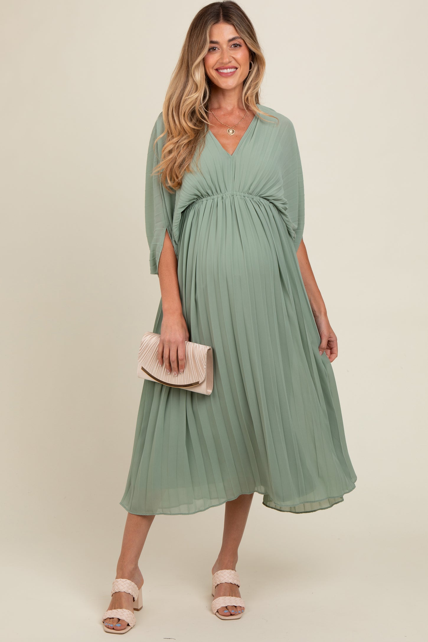 woman wearing a mint green pleated maternity dress with a v-neck and flowing sleeves, holding a clutch and showing a baby bump