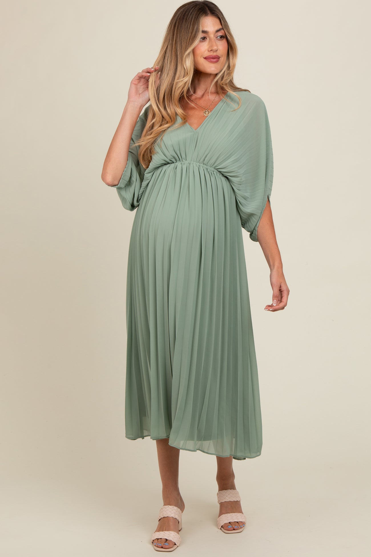 woman wearing a sage green pleated maternity dress with a v-neckline and flowy sleeves, showcasing her baby bump
