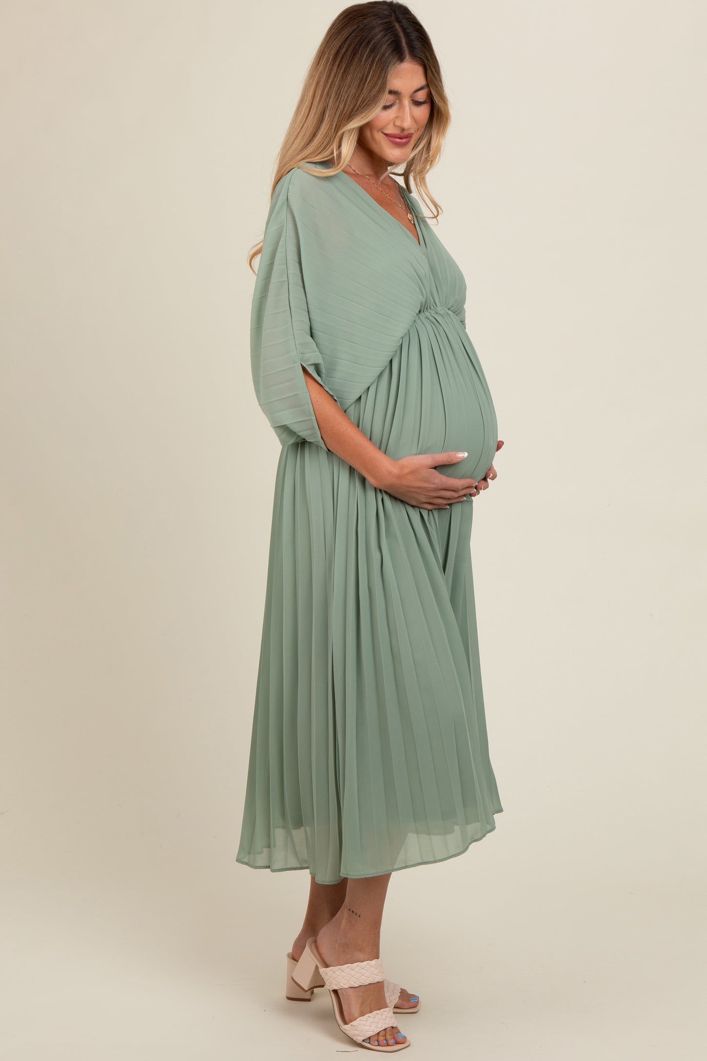 woman wearing a flowing green maternity dress with pleats, cradling her baby bump while standing sideways