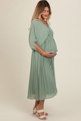 woman wearing a flowing green maternity dress with pleats, cradling her baby bump while standing sideways