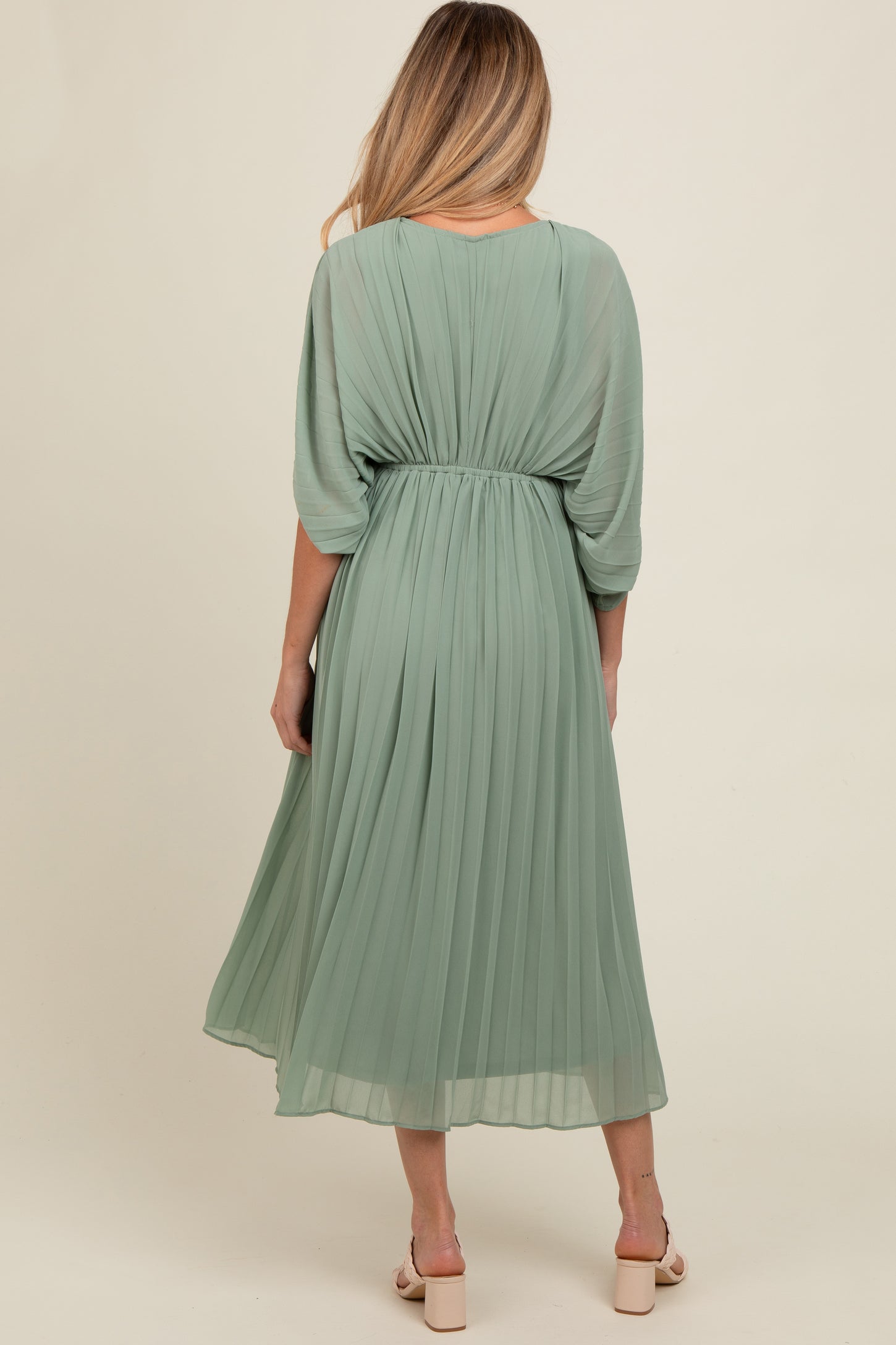 woman wearing a pleated sage green maternity dress with a gathered waist and flowy sleeves, turned away from the camera
