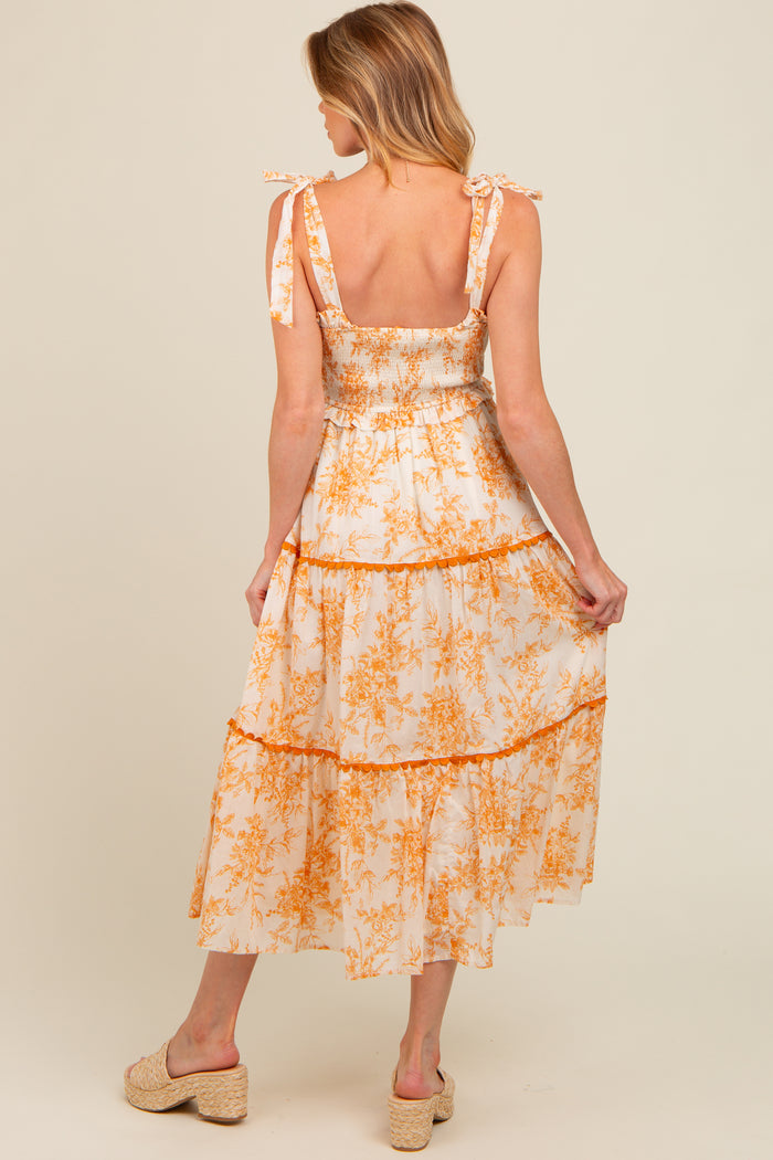 woman wearing a white and orange floral maternity dress with tie shoulder straps and a tiered skirt, showing a baby bump