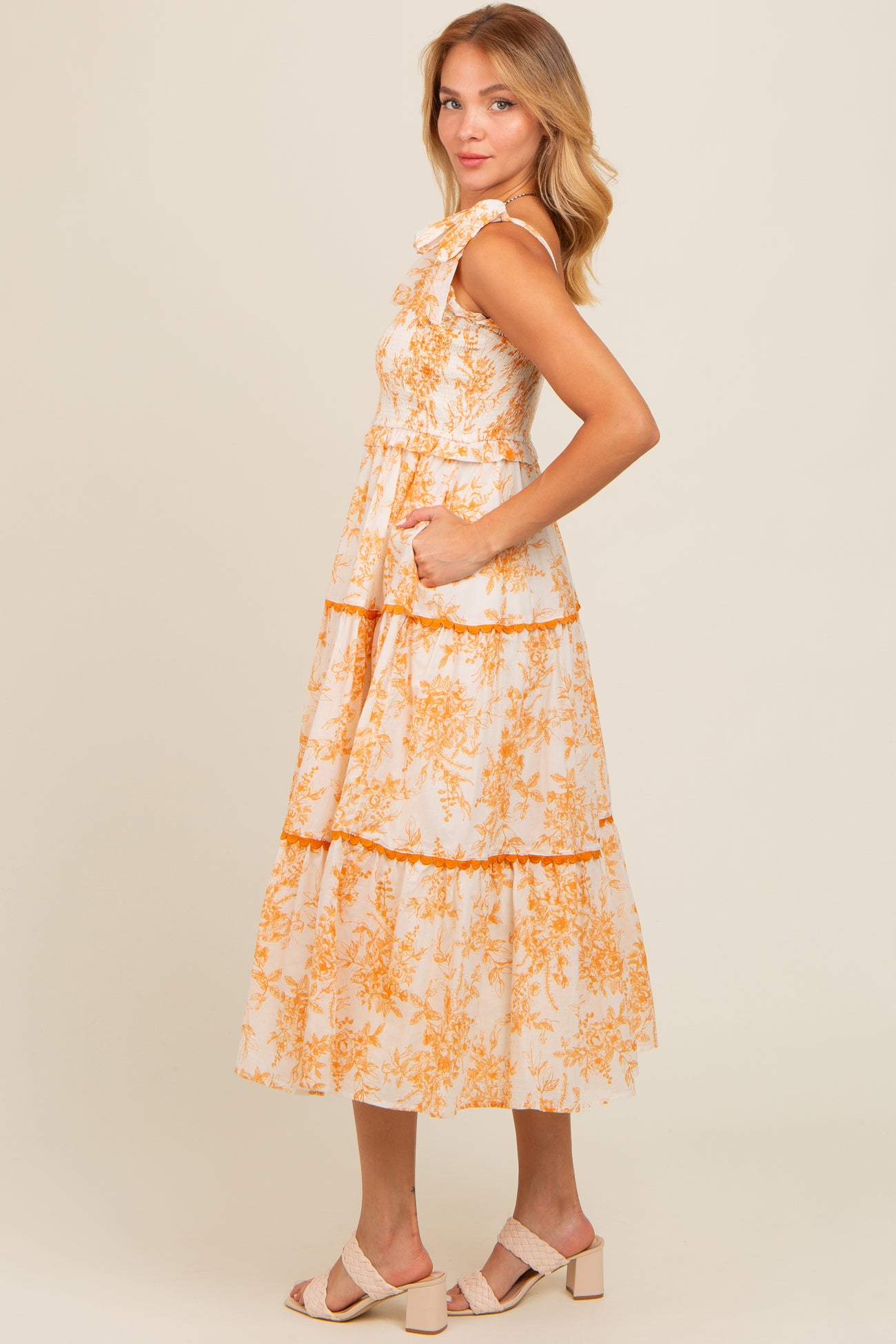 woman wearing a floral print sleeveless midi dress with tiered ruffles and pockets in peach and cream colors