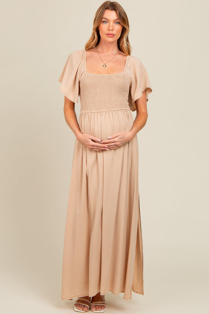 woman wearing a beige maternity maxi dress with a smocked bodice and flutter sleeves, gently holding her baby bump