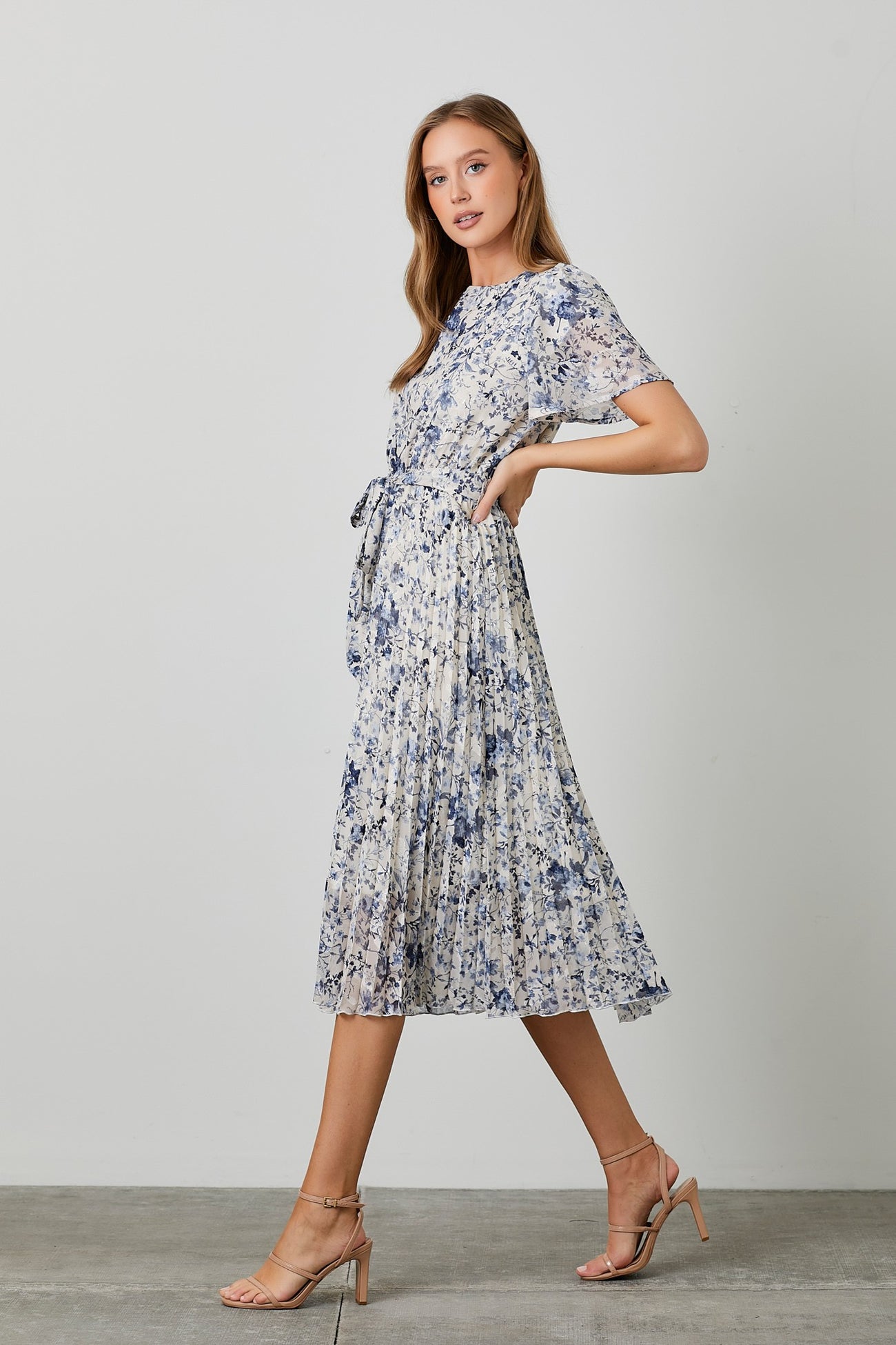 woman wearing a floral pleated midi dress with short sleeves and a tie waist, paired with strappy nude heels
