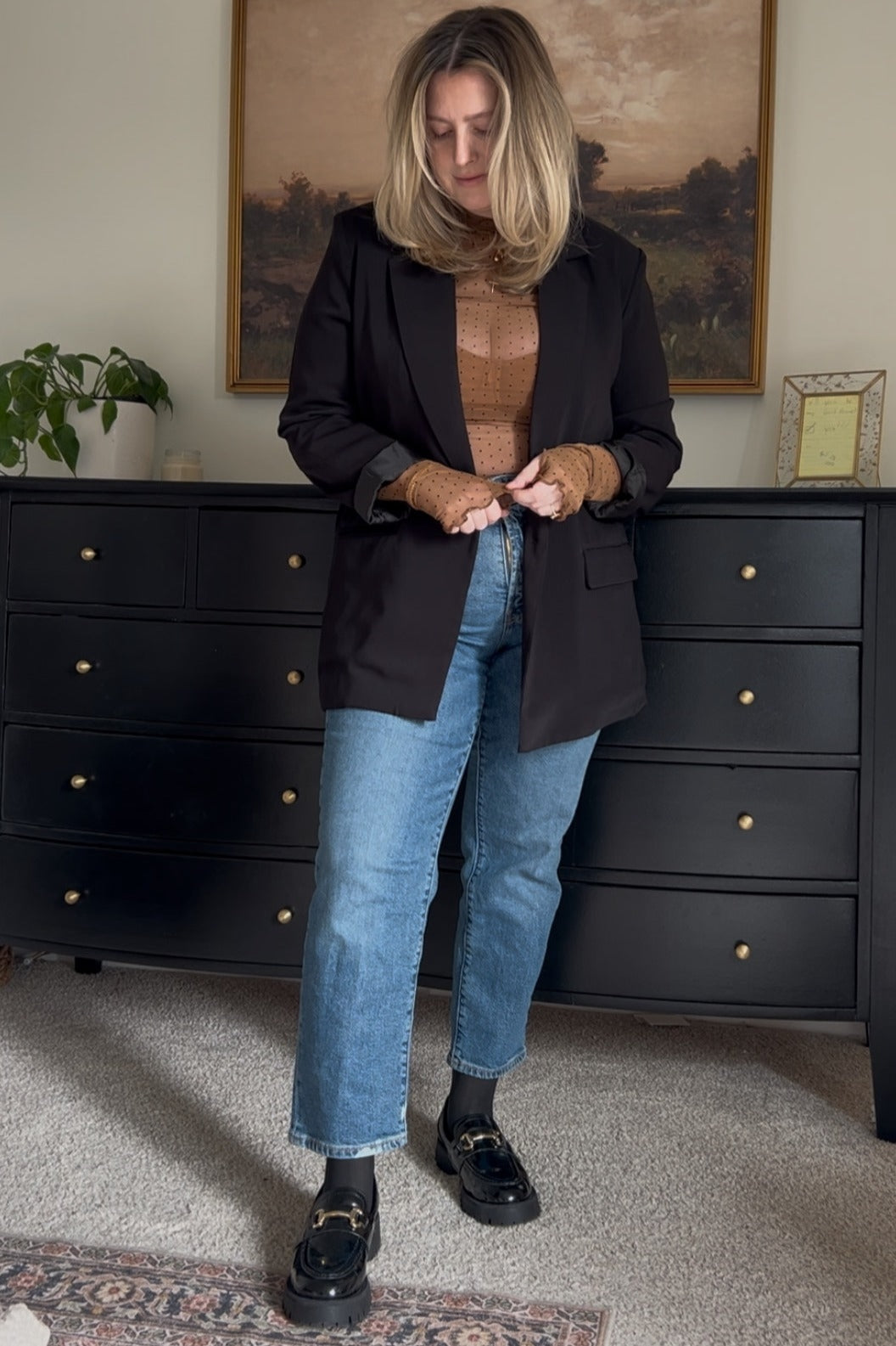 woman wearing a black blazer over a brown knit top and blue denim jeans, styled with black loafers and wavy hair