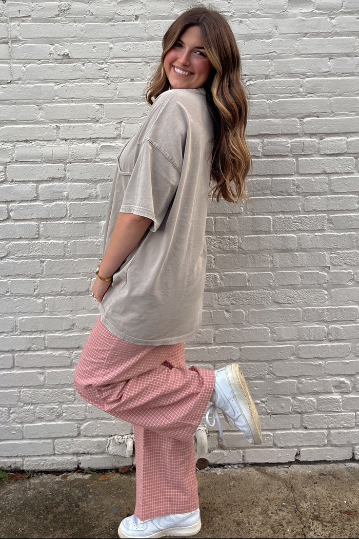 woman wearing a beige oversized t-shirt and pink checkered wide-leg pants, posing with one foot lifted on a brick background