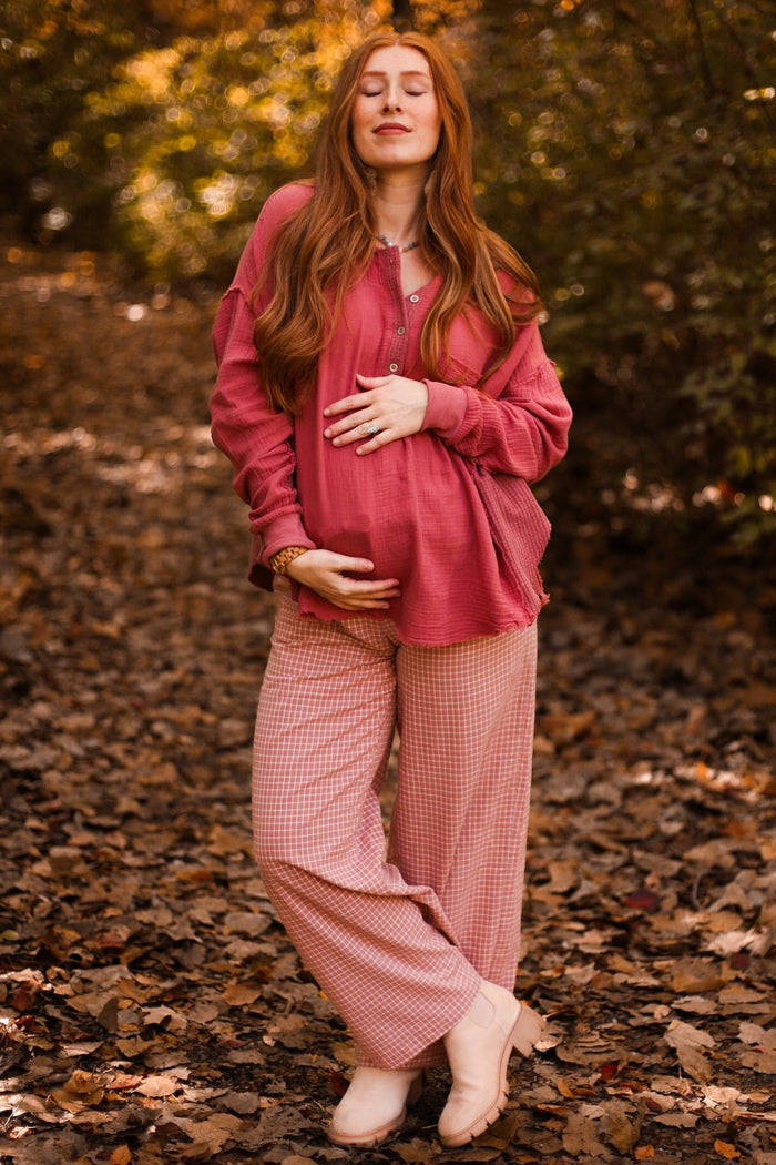 woman wearing a pink maternity top and checkered pants, holding her baby bump in an autumn setting