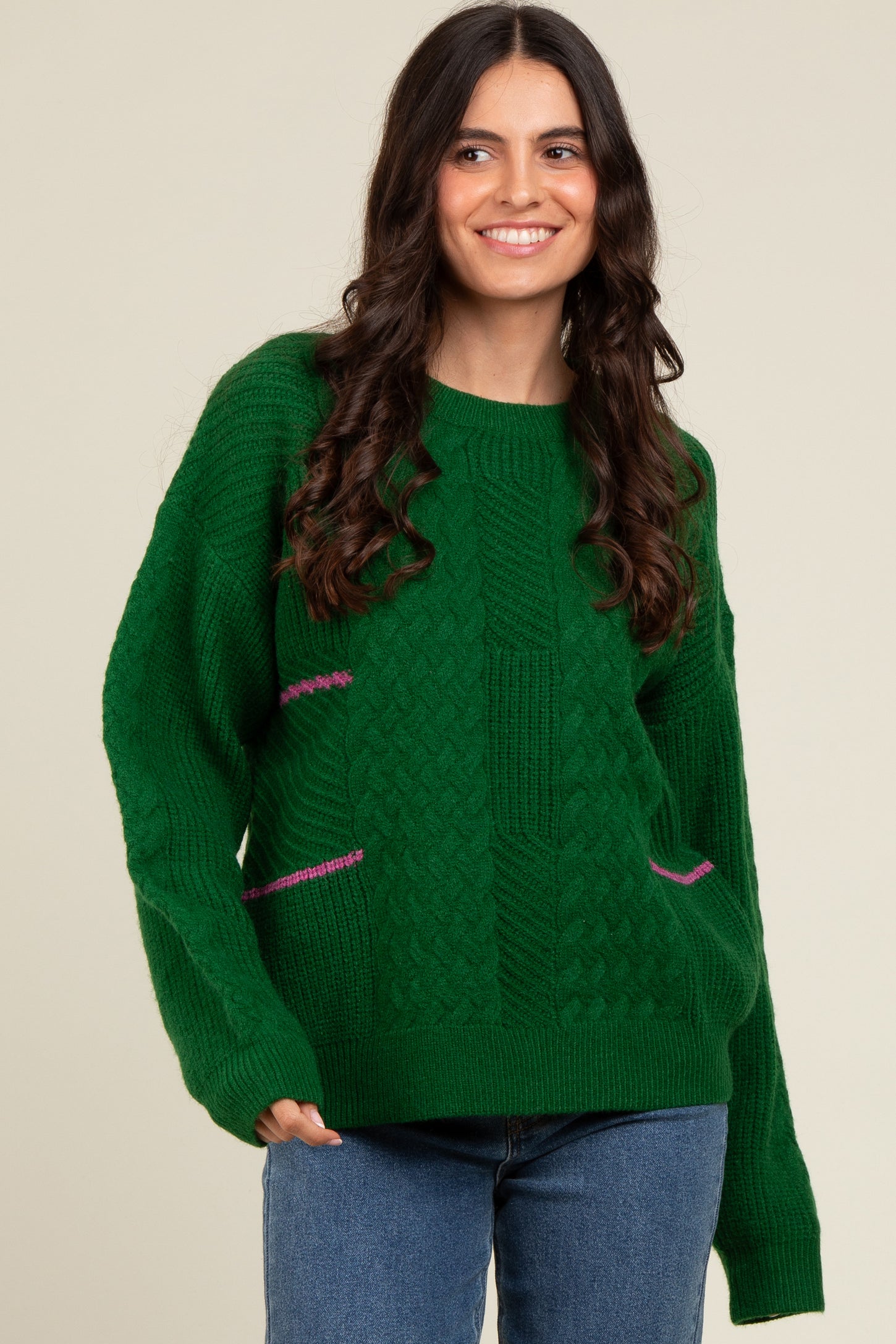 woman wearing a green cable knit sweater with pink detailing and long sleeves, smiling against a neutral backdrop