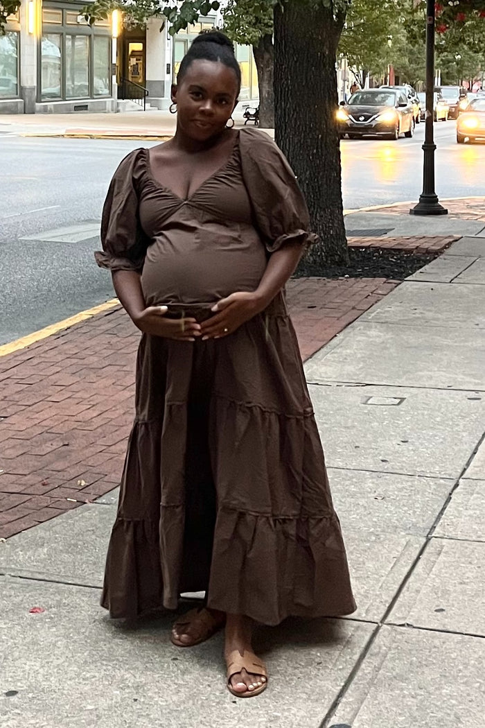 woman wearing a brown maternity maxi dress with puff sleeves and a tiered skirt, holding her baby bump outdoors