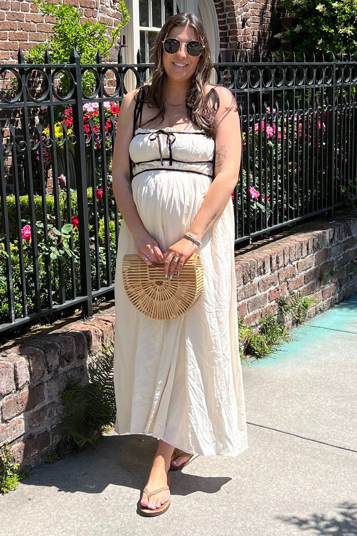 woman wearing a beige maternity maxi dress with a tie front and holding a bamboo clutch, showcasing her baby bump