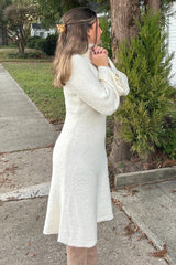 woman wearing a cream-colored long sleeve sweater dress with a flared hem standing outdoors with hands clasped