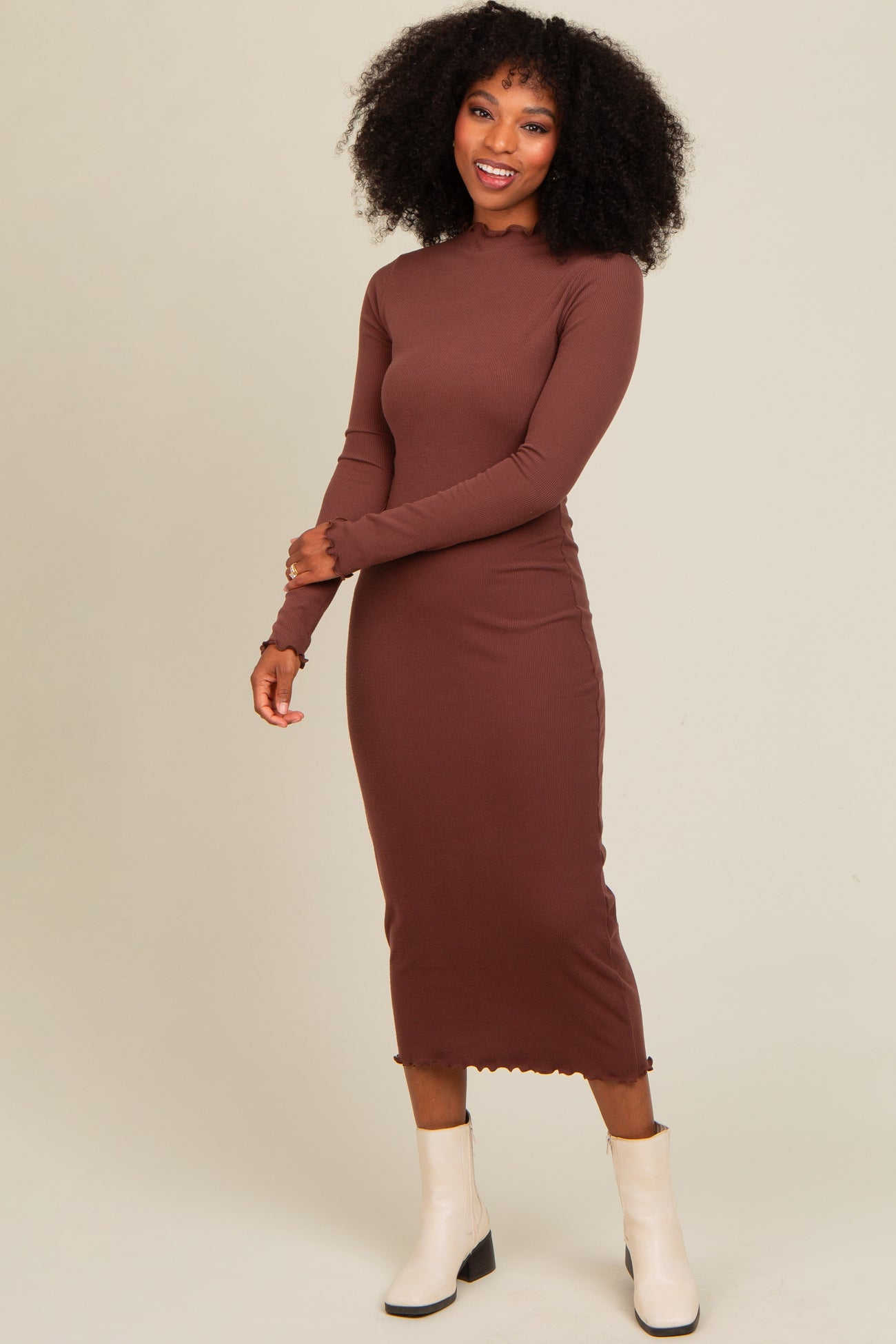 woman wearing a brown long sleeve ribbed midi dress with a fitted silhouette and a scalloped hem, paired with white ankle boots