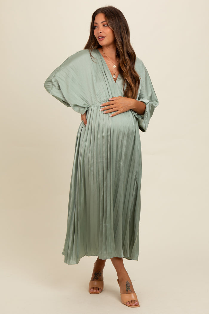 woman wearing a sage green pleated maternity dress with a deep v-neck and flowing sleeves, holding her baby bump