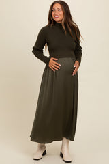 woman wearing a dark green long sleeve maternity dress and a fitted sweater, holding her baby bump