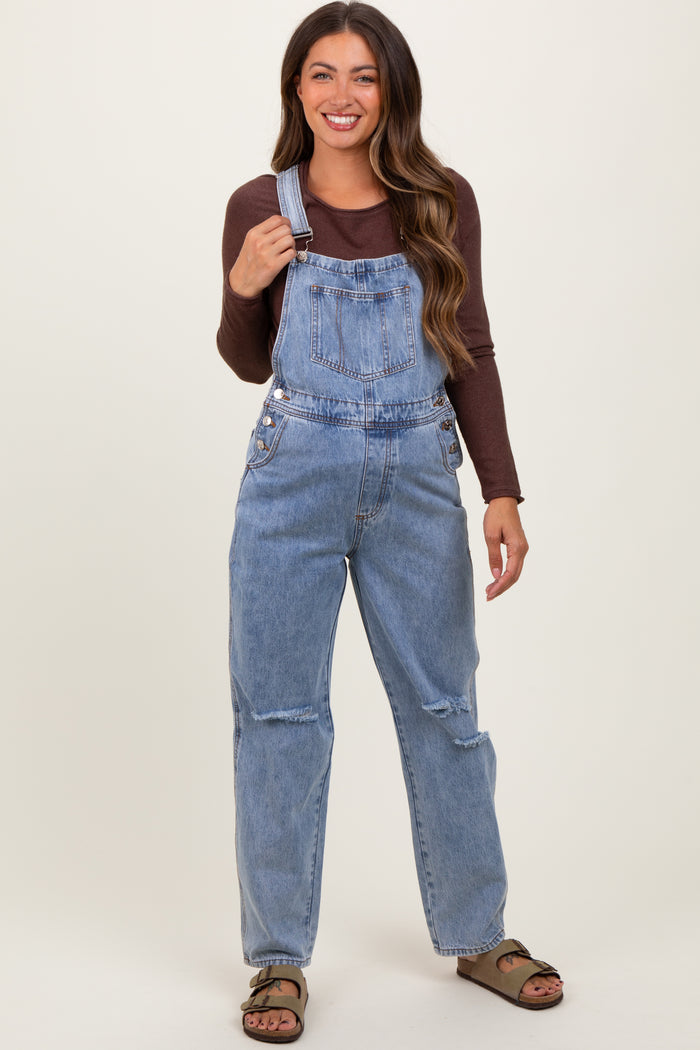 woman wearing a light blue maternity denim overall with a brown long sleeve top and sandals, holding the straps