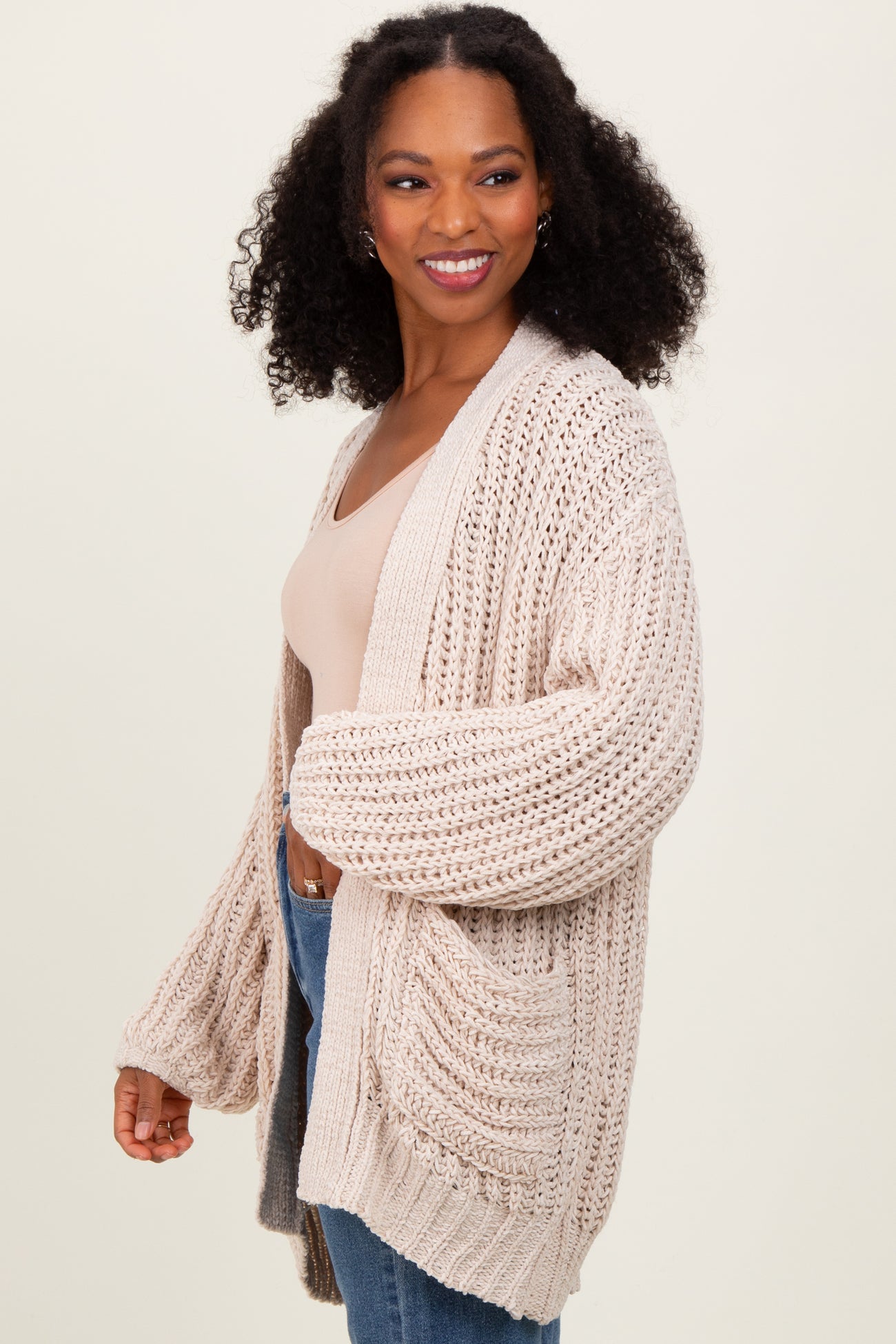 woman wearing a chunky beige knit cardigan over a light top and denim jeans, smiling and posing to the side