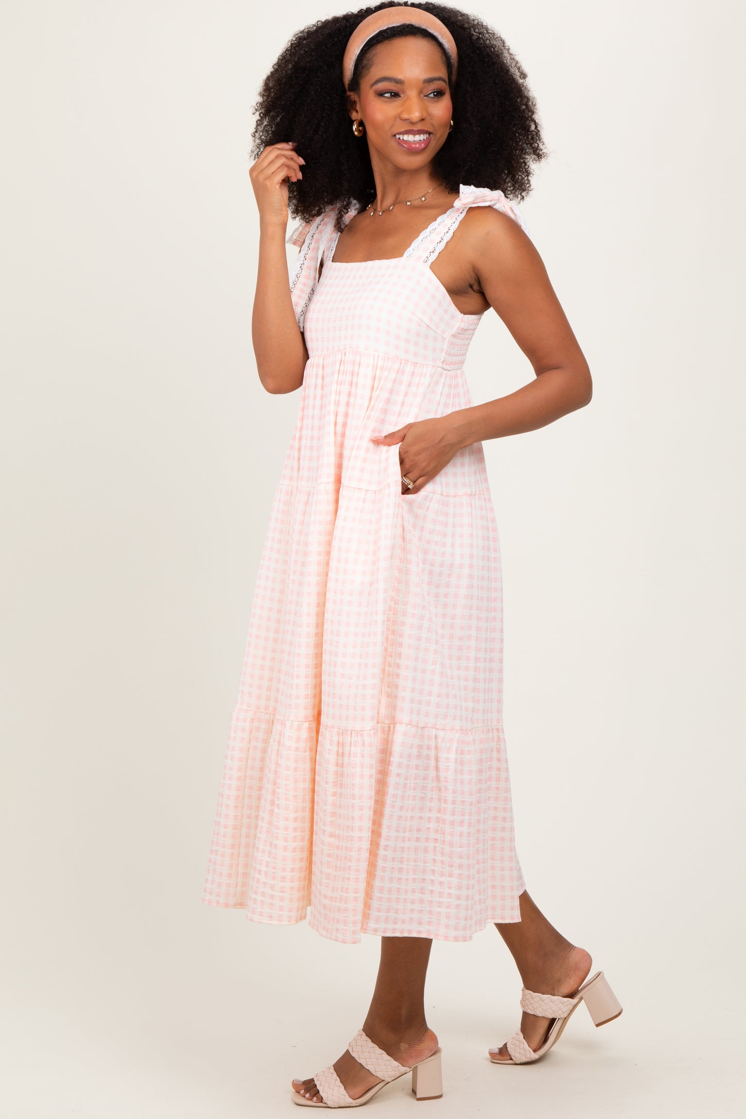 woman wearing a light pink gingham midi dress with ruffled straps and pockets, styled with a headband and sandals