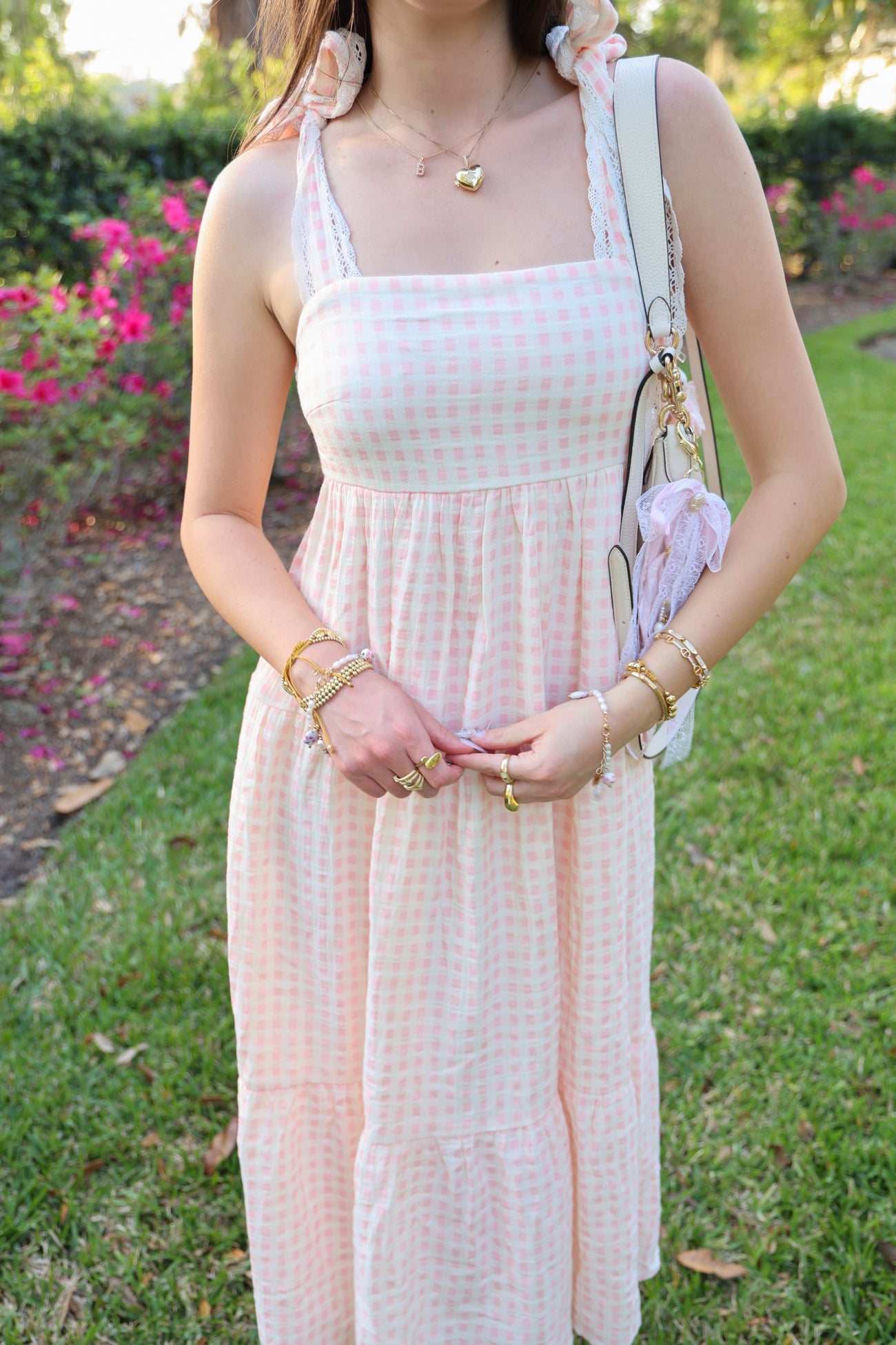 woman wearing a pink and white gingham sundress with lace straps and holding a small handbag on a grassy area