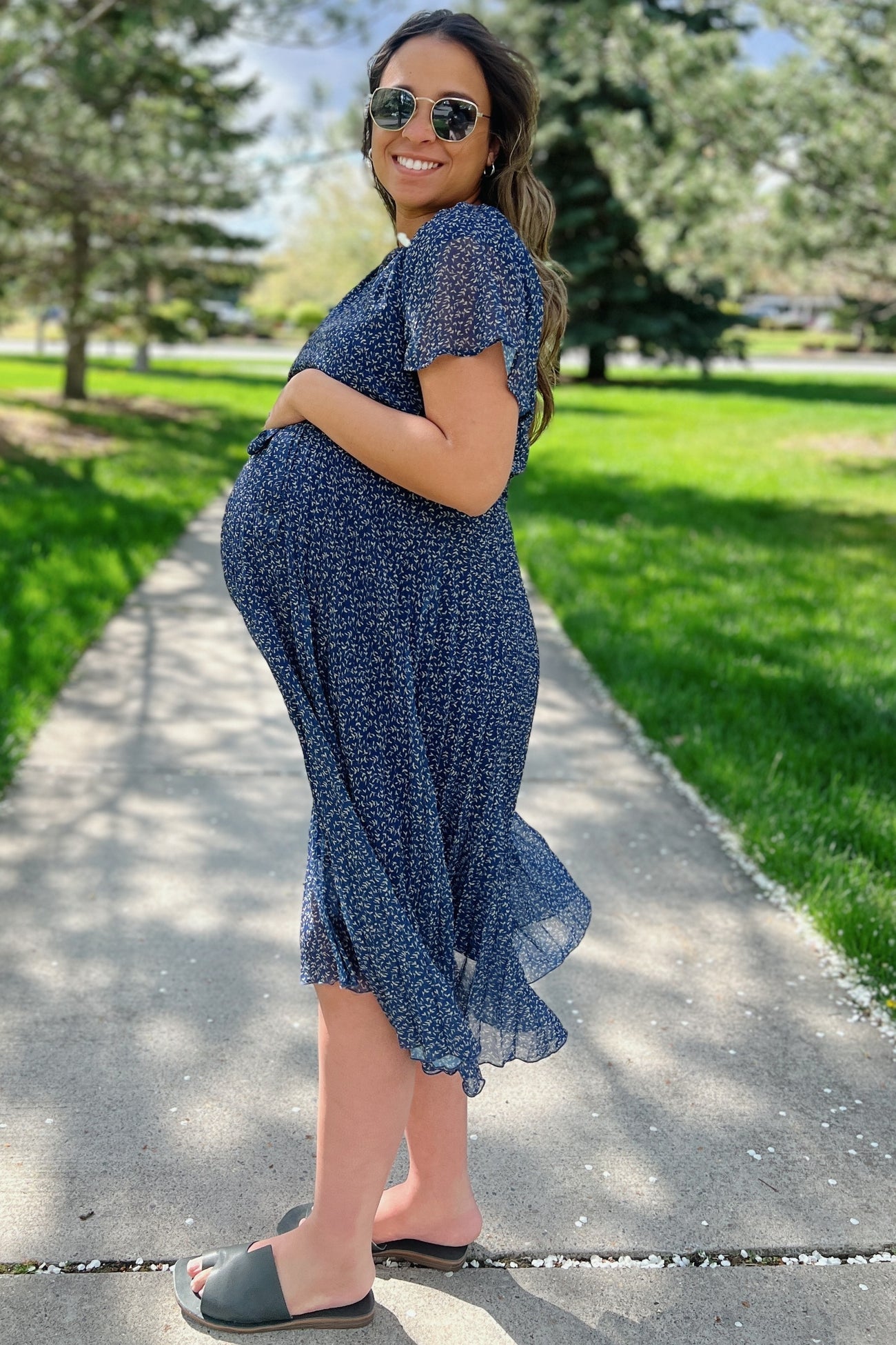 woman wearing a navy blue floral maternity dress, smiling and showing her baby bump outdoors in a park