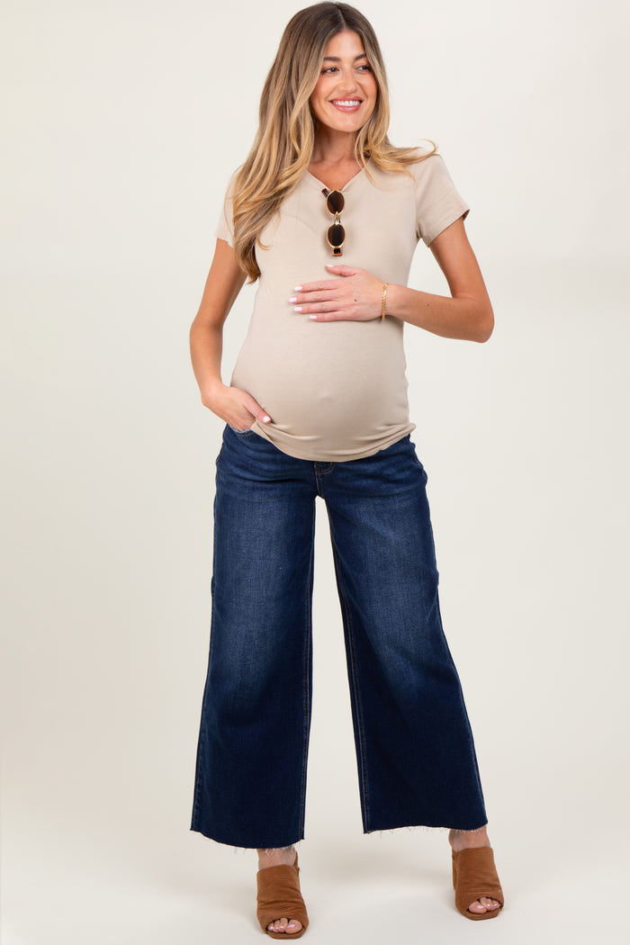 woman wearing a beige short sleeve maternity top and blue wide-leg jeans, holding her baby bump