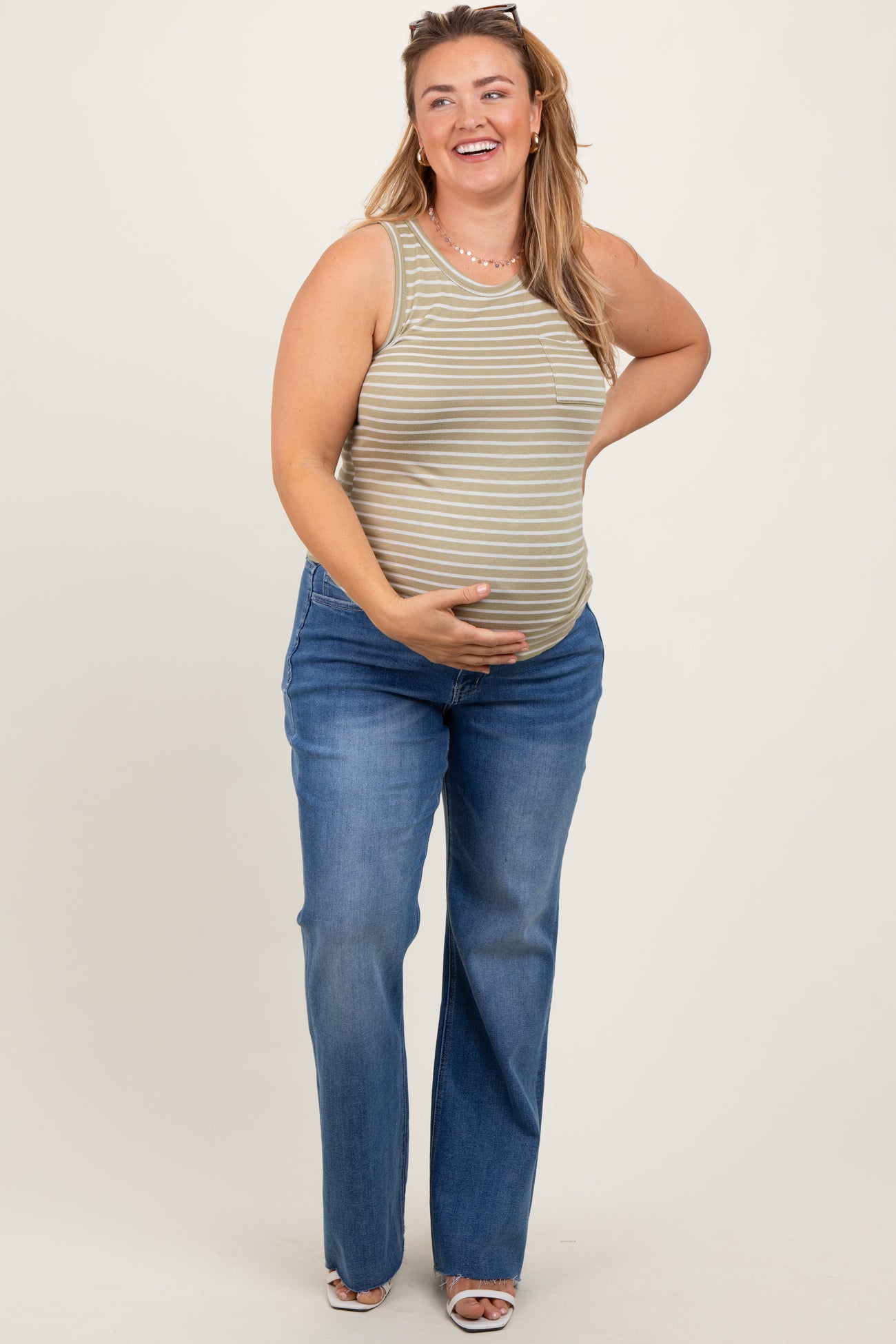 woman wearing a striped green maternity tank top and blue maternity jeans, smiling and holding her baby bump