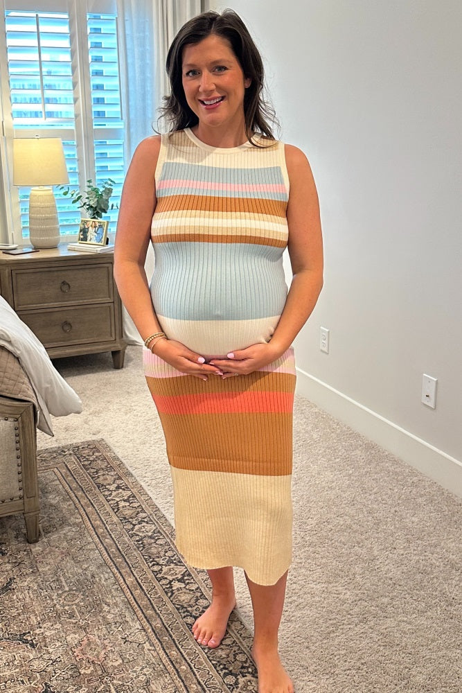 woman wearing a multicolored striped maternity dress, holding her baby bump while standing in a cozy room