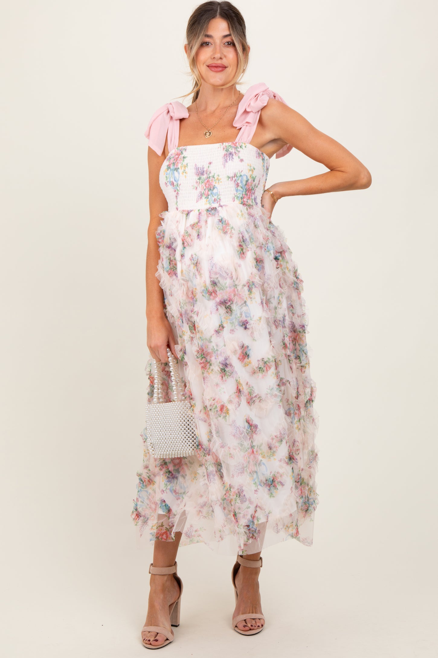 woman wearing a floral maternity dress with ruffled details and pink bow straps, holding a small bag and showing her baby bump