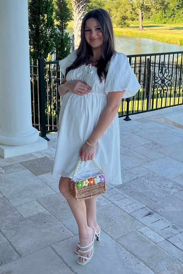 woman wearing a white maternity dress with puff sleeves, holding her baby bump and a basket of colorful eggs