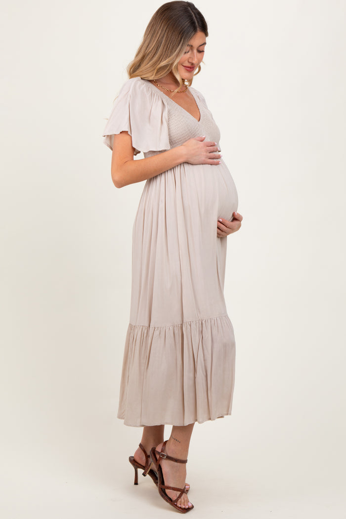 woman wearing a light beige maternity dress with flutter sleeves and a tiered hem, holding her baby bump