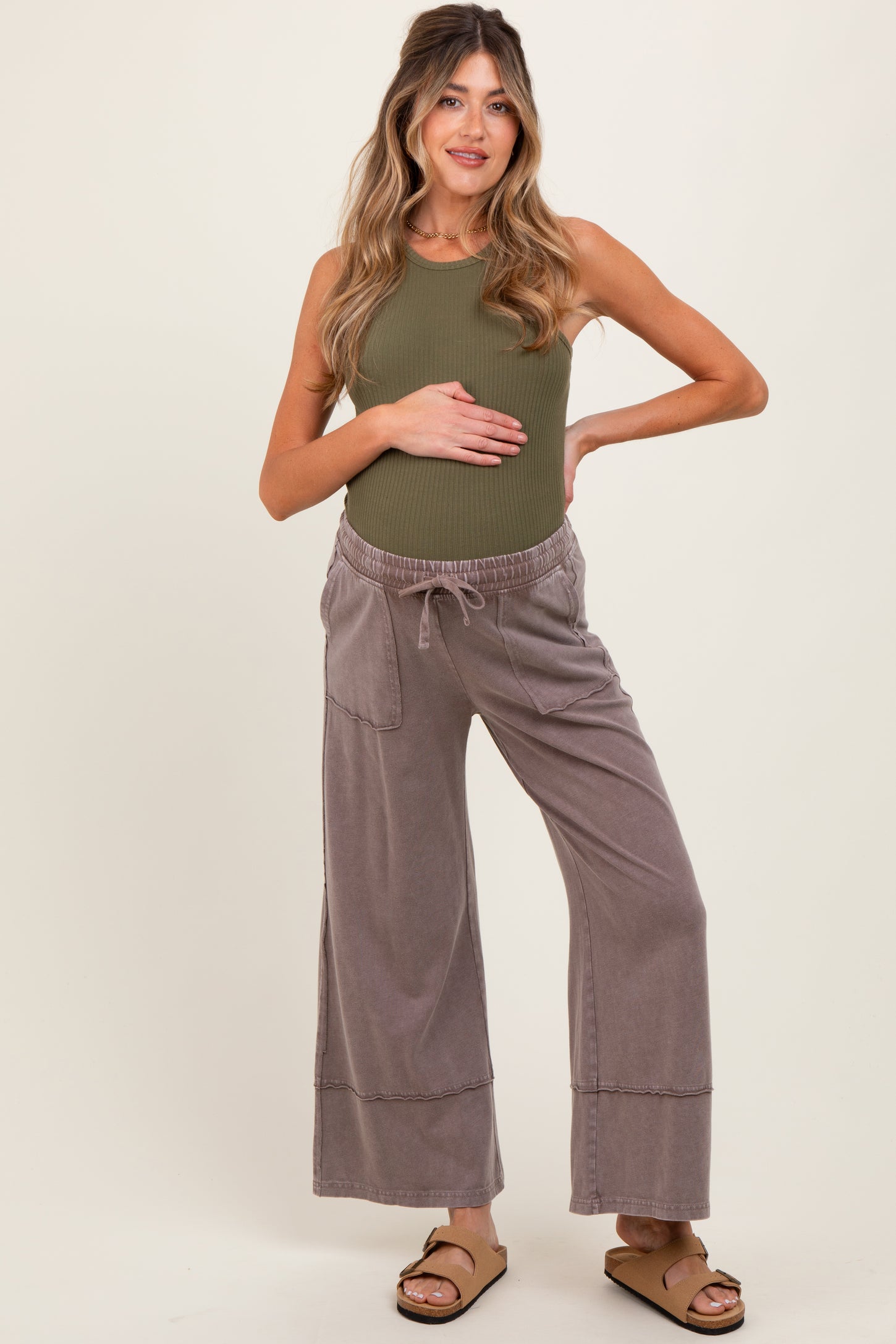 woman wearing a light olive green ribbed maternity tank top and gray wide-leg maternity pants, holding her baby bump