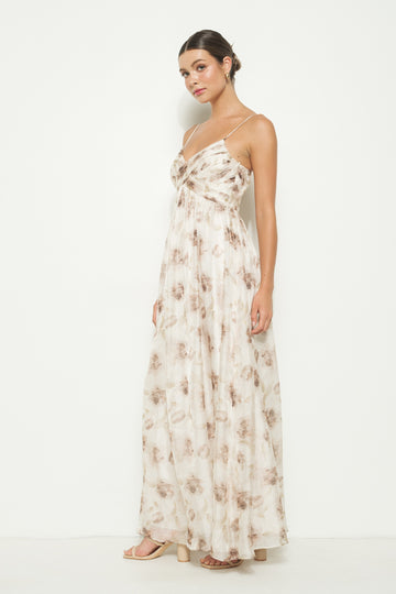 woman wearing a sleeveless maxi dress with a floral pattern in beige and brown tones, standing against a white background