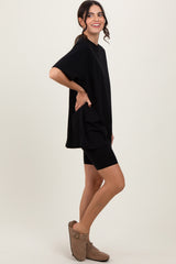 woman wearing an oversized black t-shirt and black biker shorts with brown slip-on shoes, standing sideways with a smile