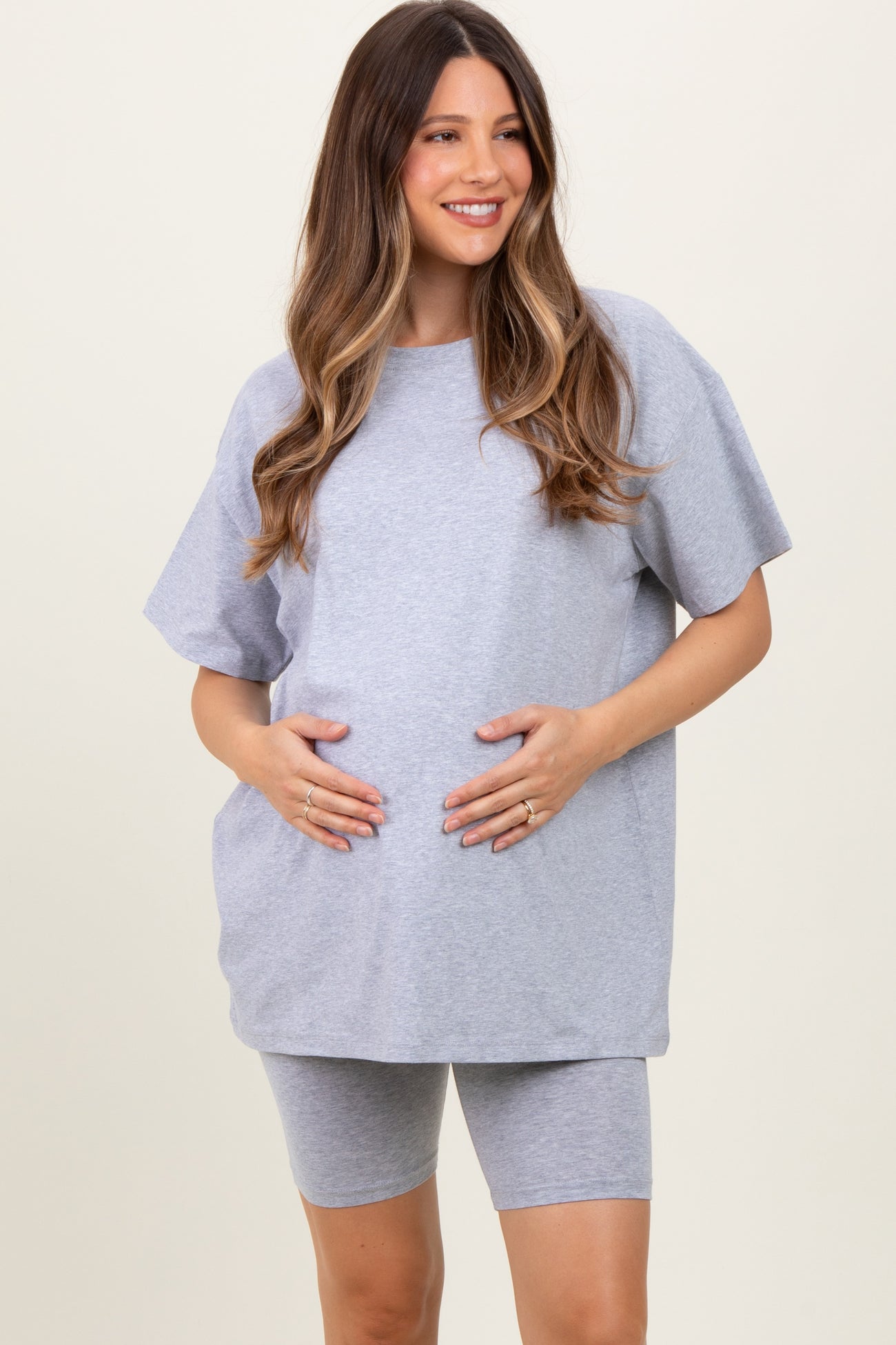 woman wearing a gray maternity t-shirt and matching shorts, holding her baby bump with a smile